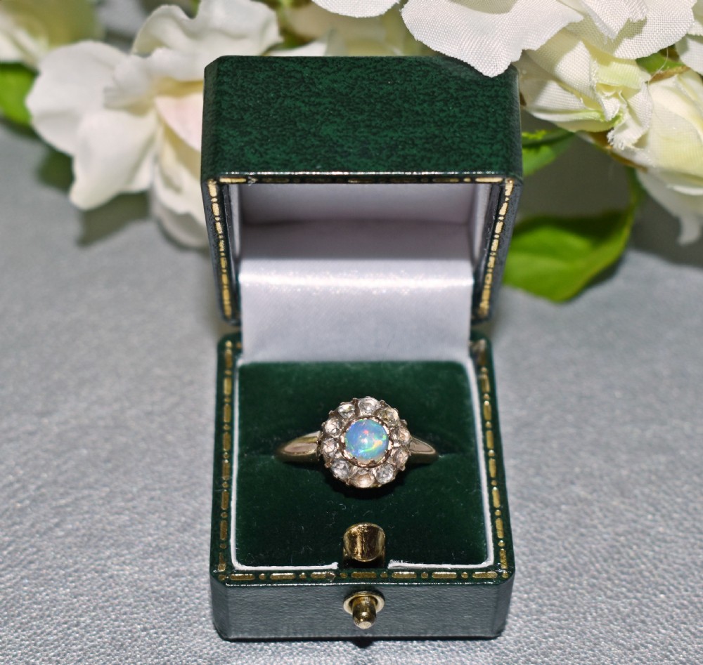 An antique gold Victorian cluster ring featuring a central cabochon opal surrounded by a halo of rose-cut diamonds, displayed inside a green velvet ring box. View VII