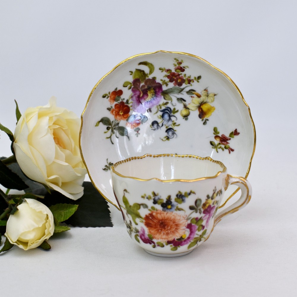 A white porcelain demitasse cup and matching saucer painted with polychrome floral sprays and finished with gold gilded scalloped edges.
