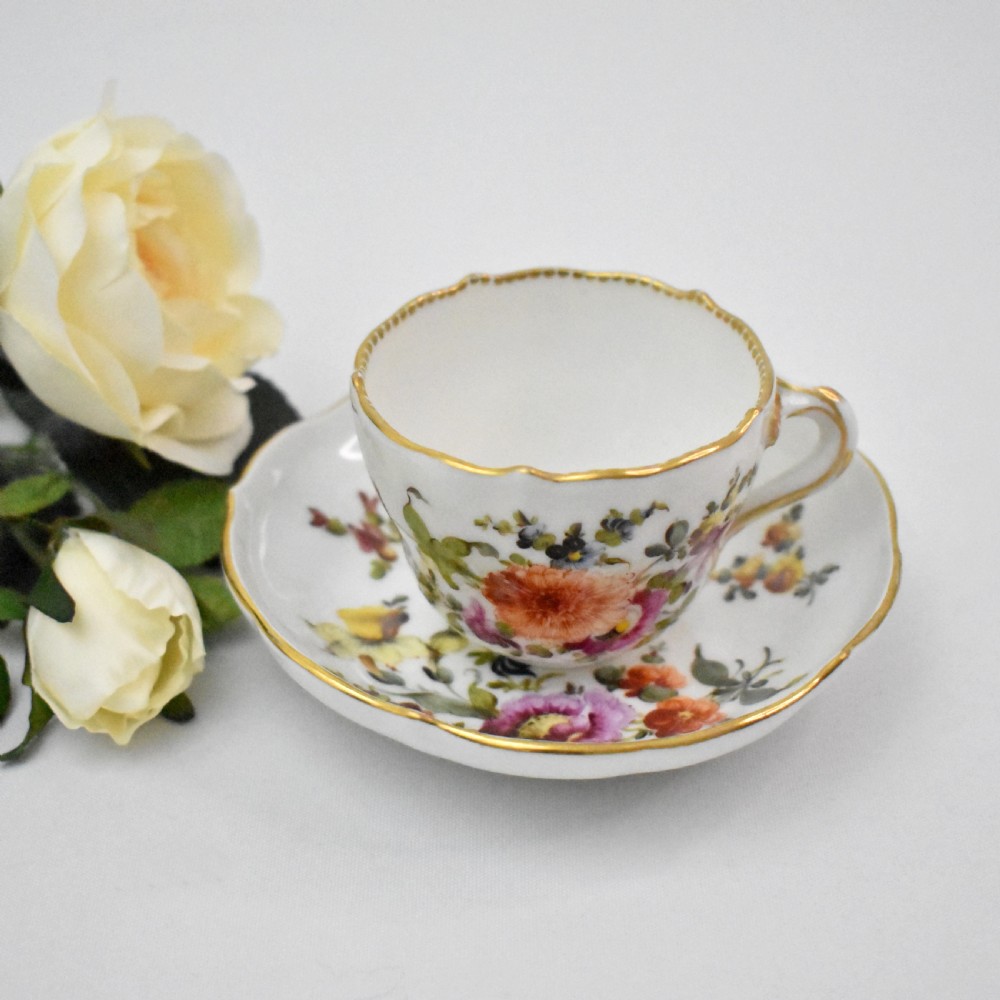 A white porcelain demitasse cup and matching saucer painted with polychrome floral sprays and finished with gold gilded scalloped edges. View II