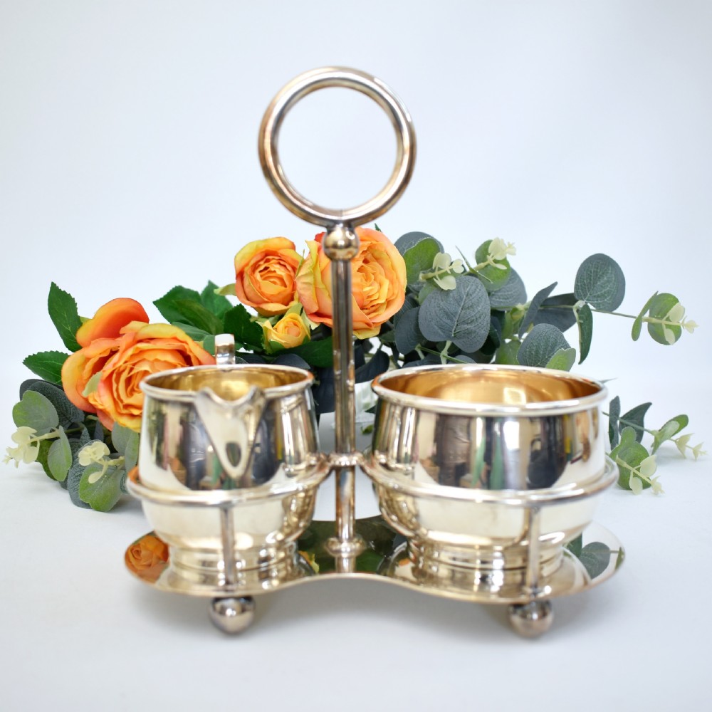 An Edwardian silver-plated cream jug and sugar bowl set held within a fitted stand featuring a central ring handle and ball feet.