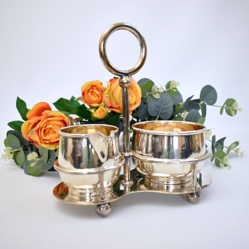 An Edwardian silver-plated cream jug and sugar bowl set held within a fitted stand featuring a central ring handle and ball feet. View II