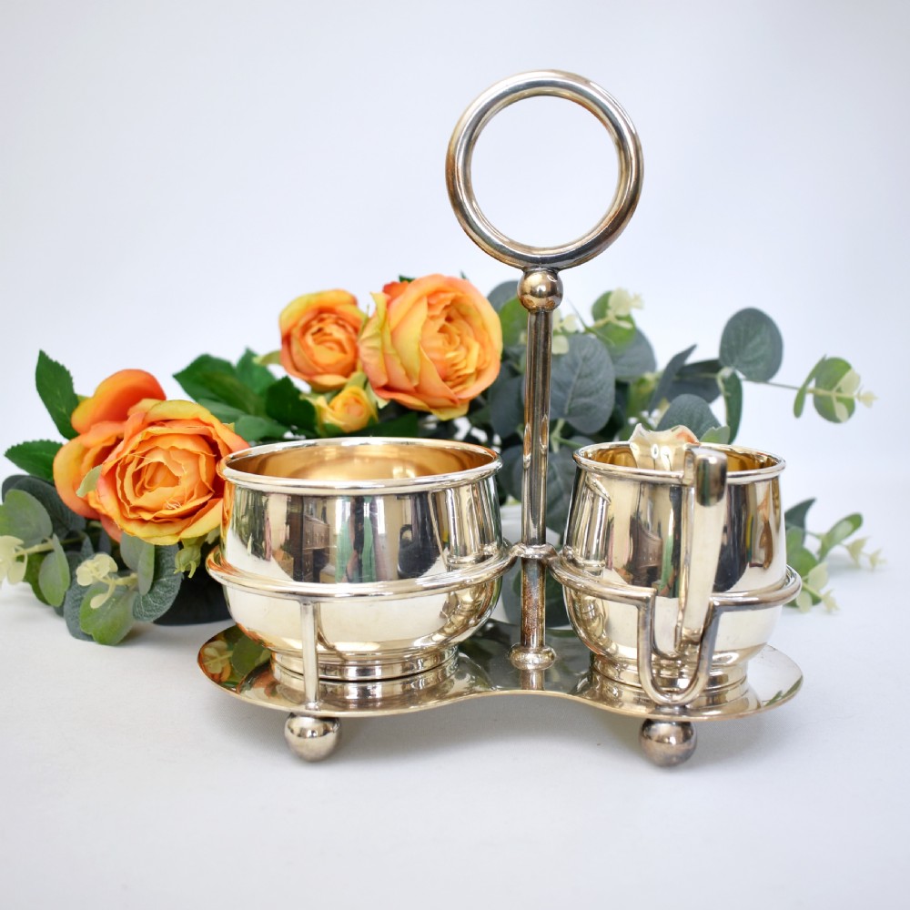 An Edwardian silver-plated cream jug and sugar bowl set held within a fitted stand featuring a central ring handle and ball feet. View V