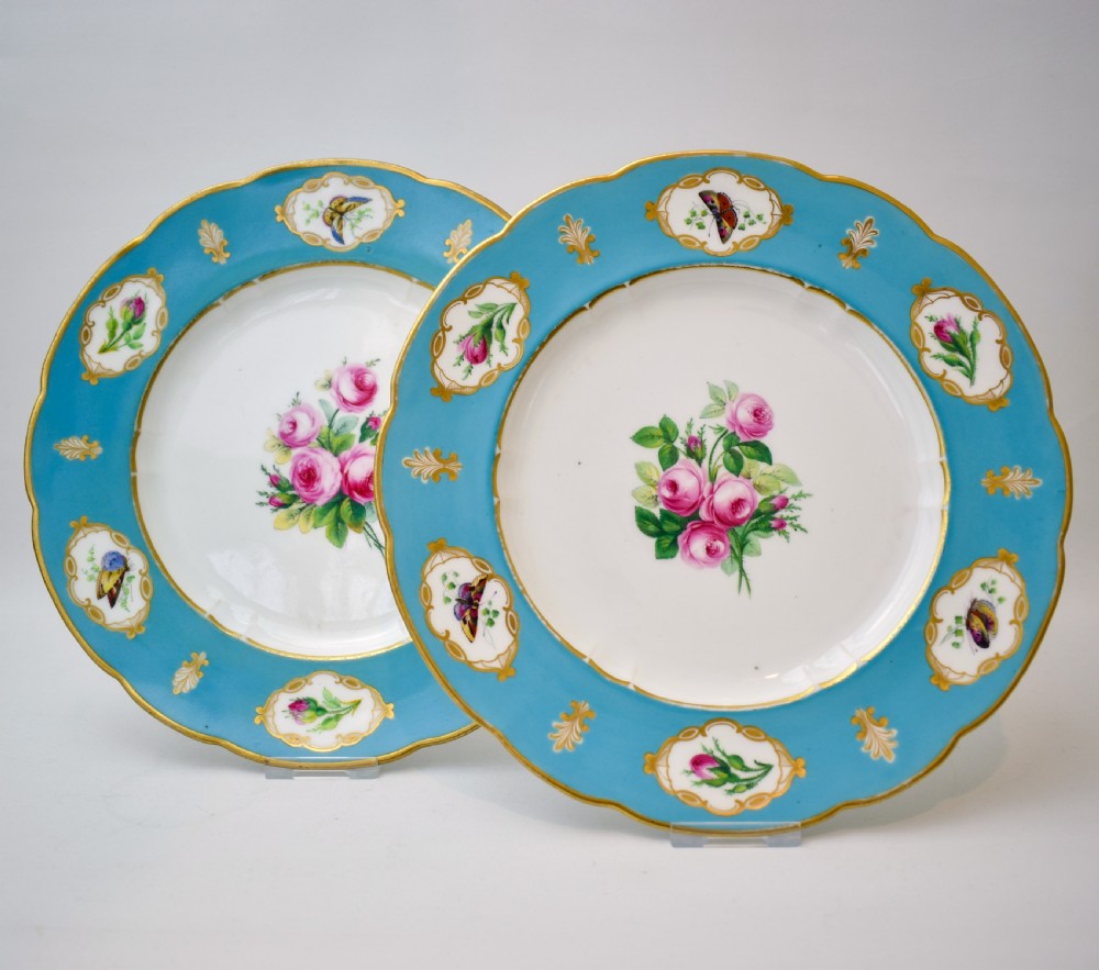 A pair of circular Victorian porcelain dessert plates featuring vibrant turquoise borders with gilded cartouches containing painted flowers and butterflies, with a central spray of pink roses on a white field.