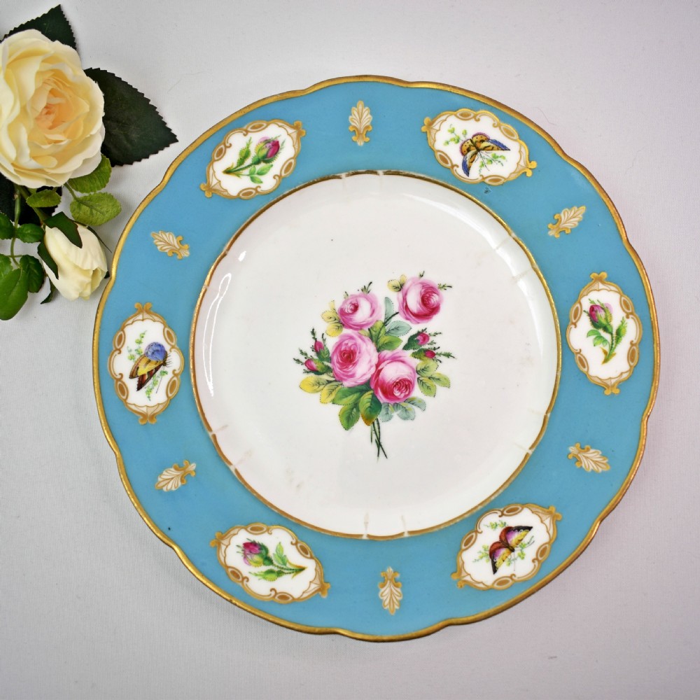 A pair of circular Victorian porcelain dessert plates featuring vibrant turquoise borders with gilded cartouches containing painted flowers and butterflies, with a central spray of pink roses on a white field. View II