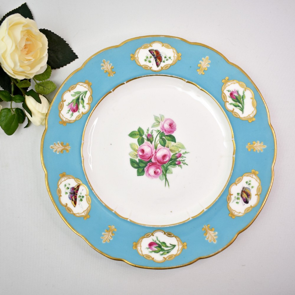 A pair of circular Victorian porcelain dessert plates featuring vibrant turquoise borders with gilded cartouches containing painted flowers and butterflies, with a central spray of pink roses on a white field. View VI