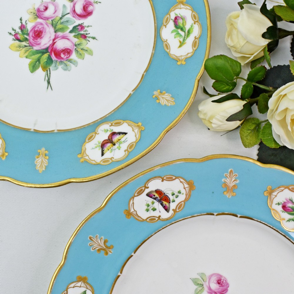 A pair of circular Victorian porcelain dessert plates featuring vibrant turquoise borders with gilded cartouches containing painted flowers and butterflies, with a central spray of pink roses on a white field. View VII