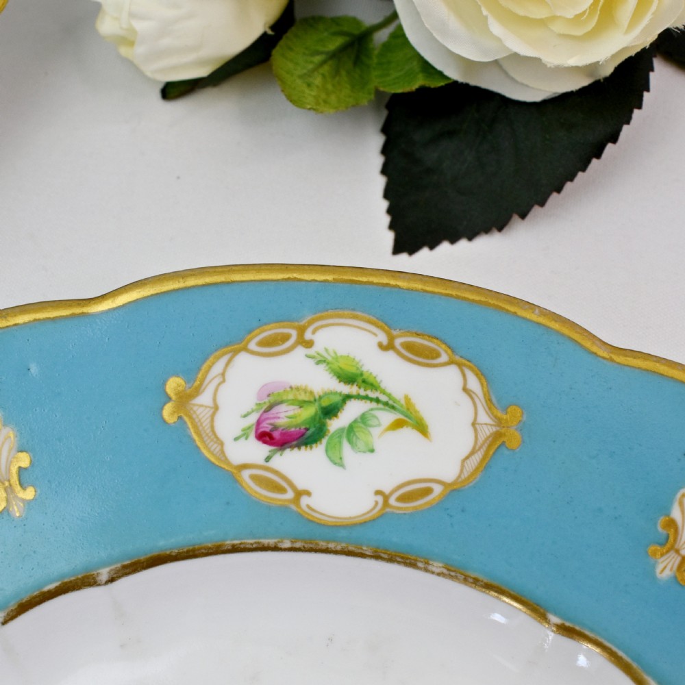 A pair of circular Victorian porcelain dessert plates featuring vibrant turquoise borders with gilded cartouches containing painted flowers and butterflies, with a central spray of pink roses on a white field. View X