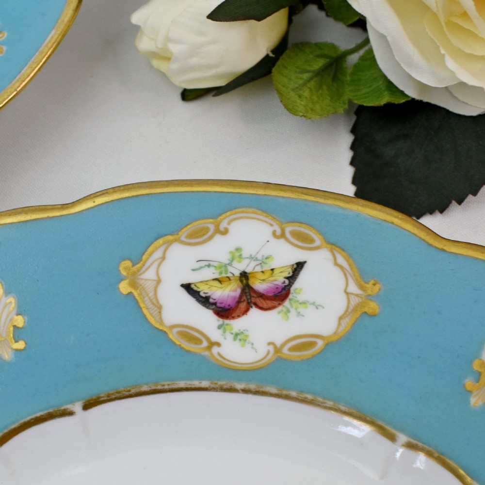 A pair of circular Victorian porcelain dessert plates featuring vibrant turquoise borders with gilded cartouches containing painted flowers and butterflies, with a central spray of pink roses on a white field. View XI