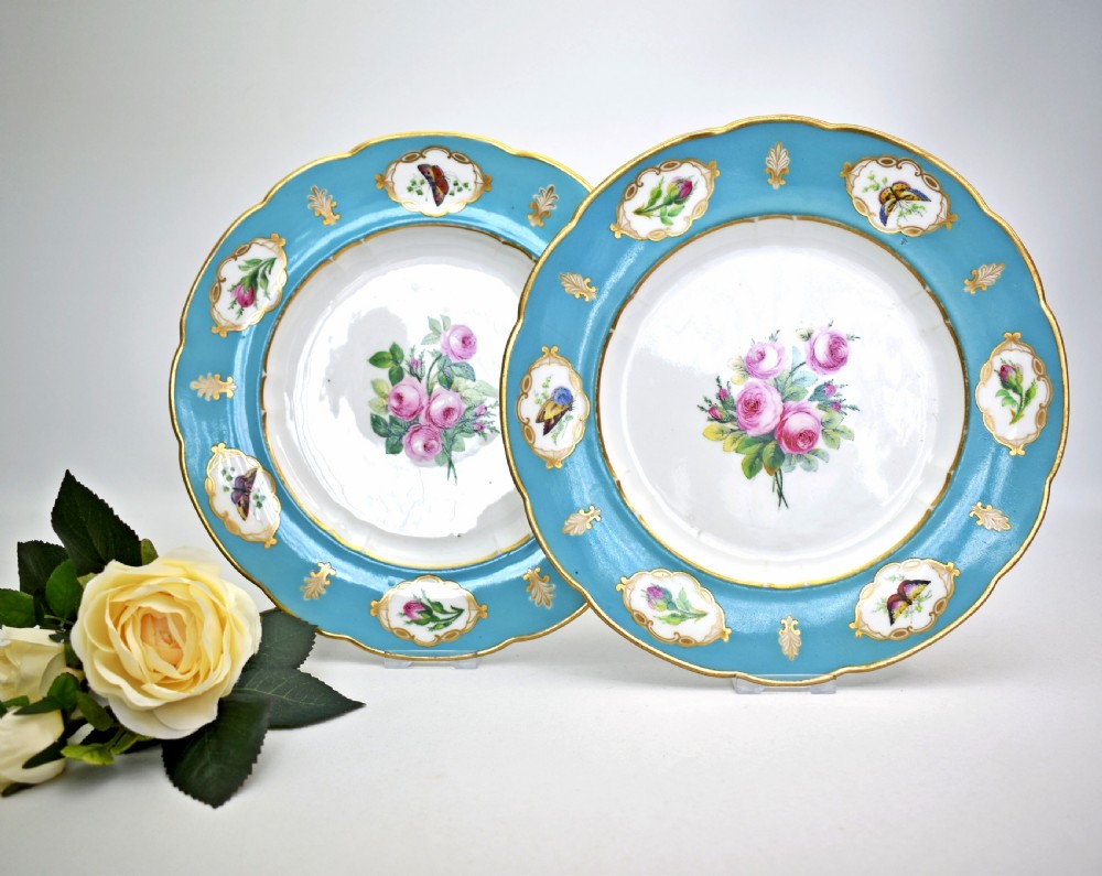 A pair of circular Victorian porcelain dessert plates featuring vibrant turquoise borders with gilded cartouches containing painted flowers and butterflies, with a central spray of pink roses on a white field. View XIV