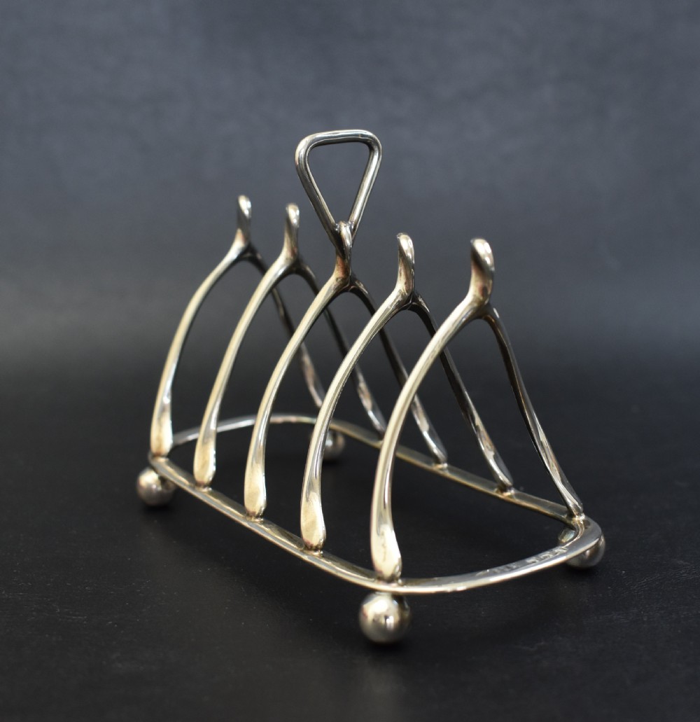 An antique sterling silver four-slot toast rack featuring wishbone-shaped dividers, a central loop handle, and ball feet. View VII