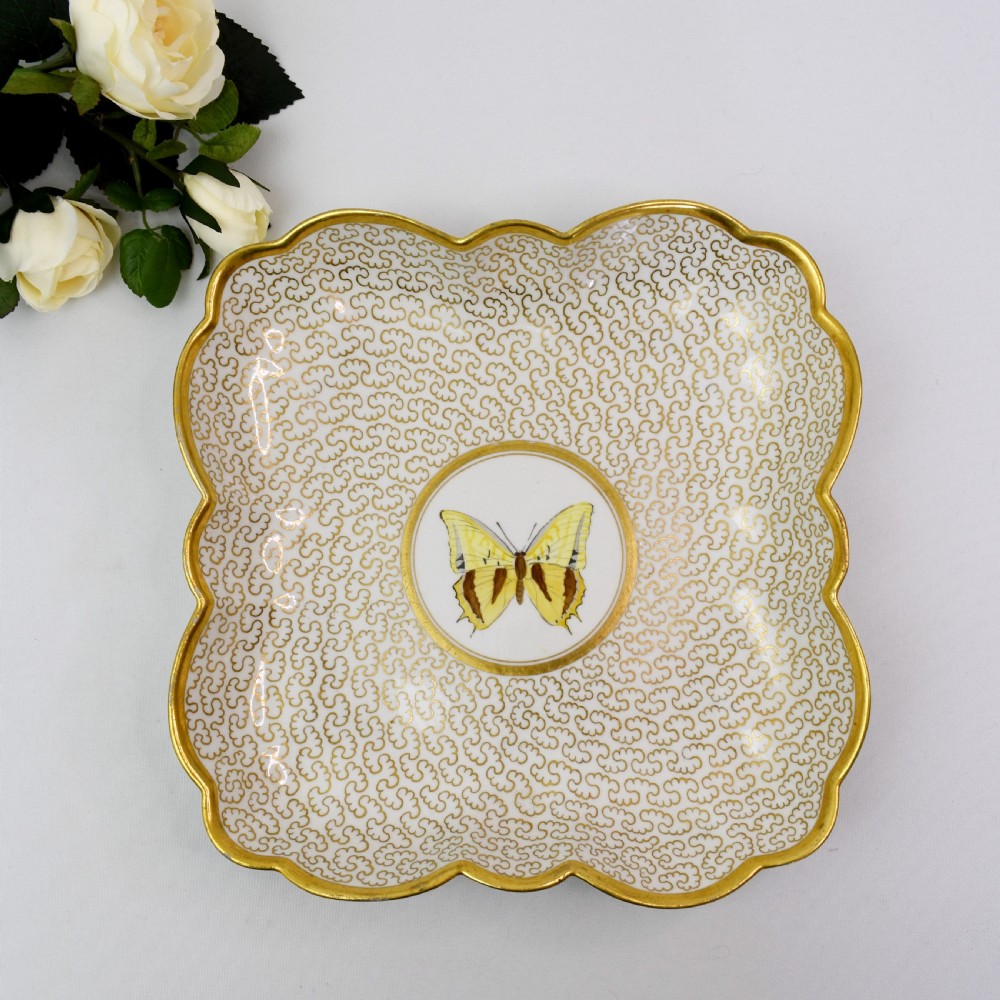 A square lobed white porcelain dish decorated with intricate gold scrollwork around a central hand-painted yellow butterfly medallion.