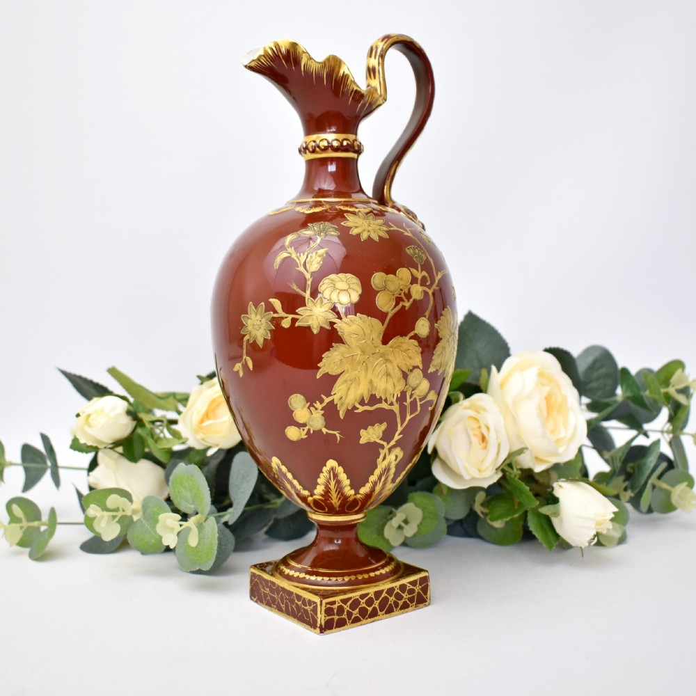 A Victorian Royal Crown Derby porcelain ewer with a terracotta brown ground, single loop handle, flared spout, and raised gold floral gilding on a square pedestal base.