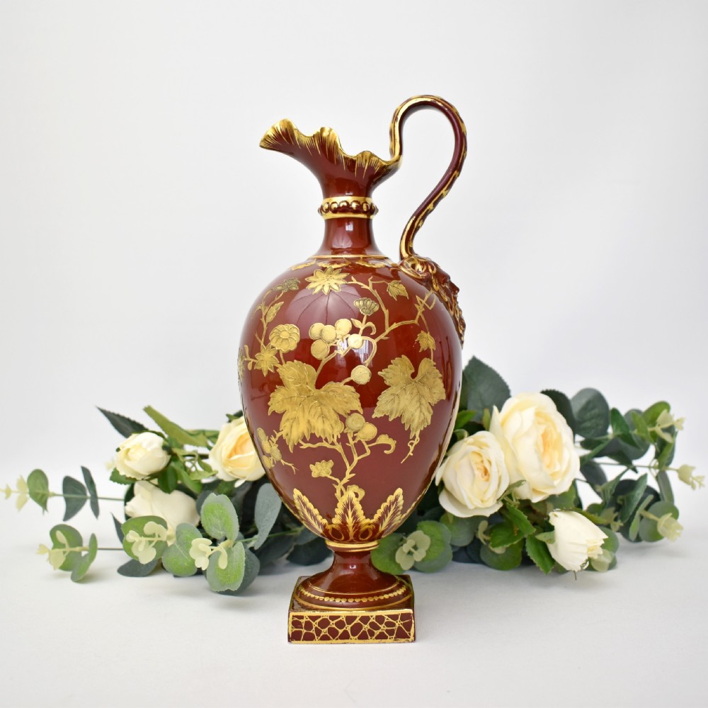 A Victorian Royal Crown Derby porcelain ewer with a terracotta brown ground, single loop handle, flared spout, and raised gold floral gilding on a square pedestal base. View II