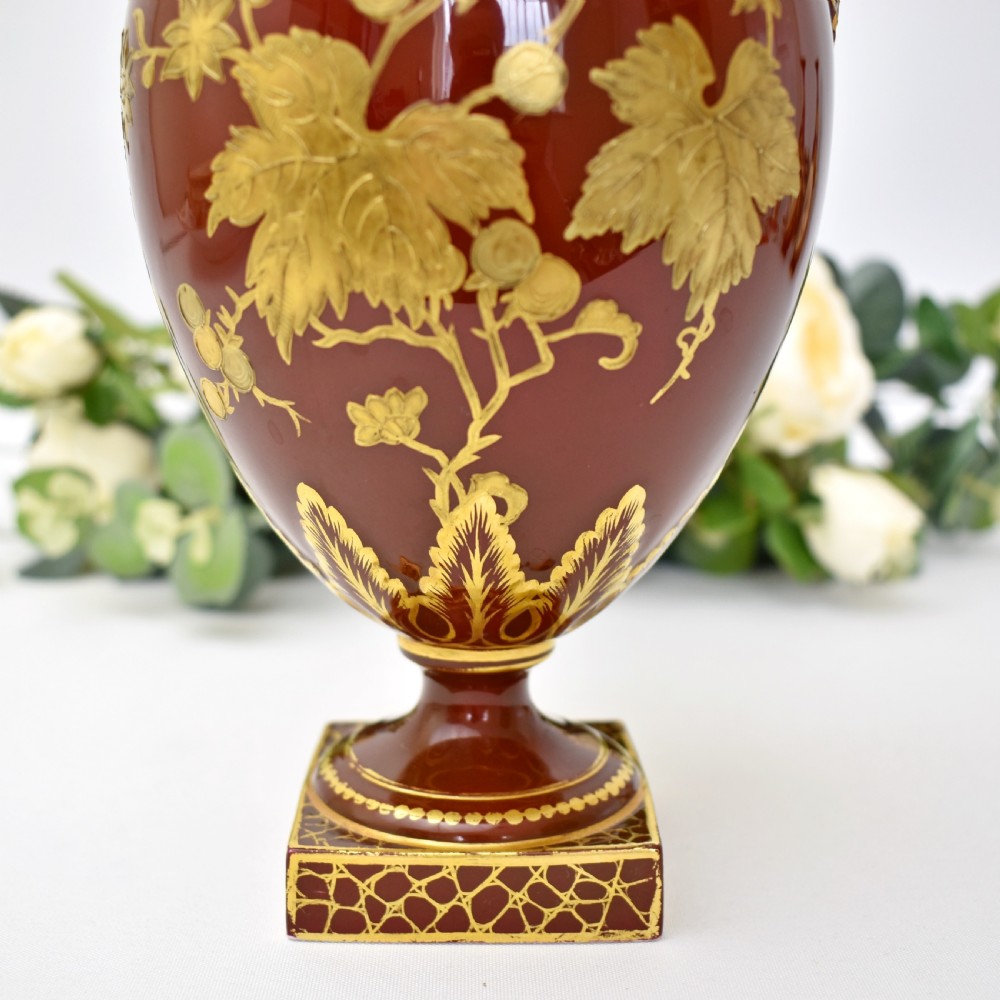A Victorian Royal Crown Derby porcelain ewer with a terracotta brown ground, single loop handle, flared spout, and raised gold floral gilding on a square pedestal base. View VI
