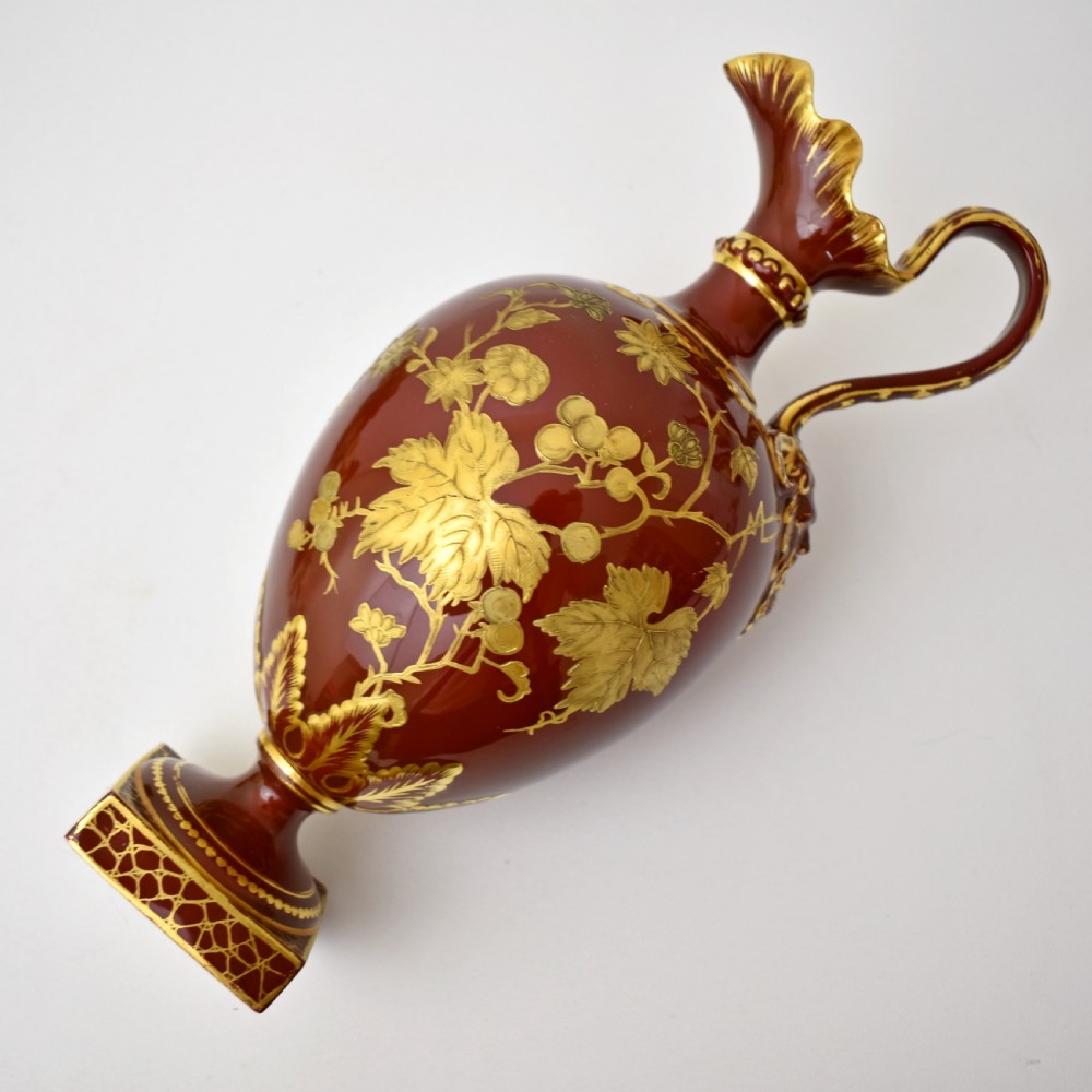 A Victorian Royal Crown Derby porcelain ewer with a terracotta brown ground, single loop handle, flared spout, and raised gold floral gilding on a square pedestal base. View X