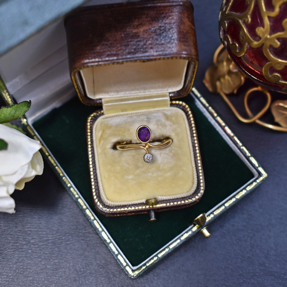 An Art Nouveau 18ct yellow gold ring featuring an oval bezel-set purple amethyst gemstone with a sweeping band and a small drop-set diamond accent.