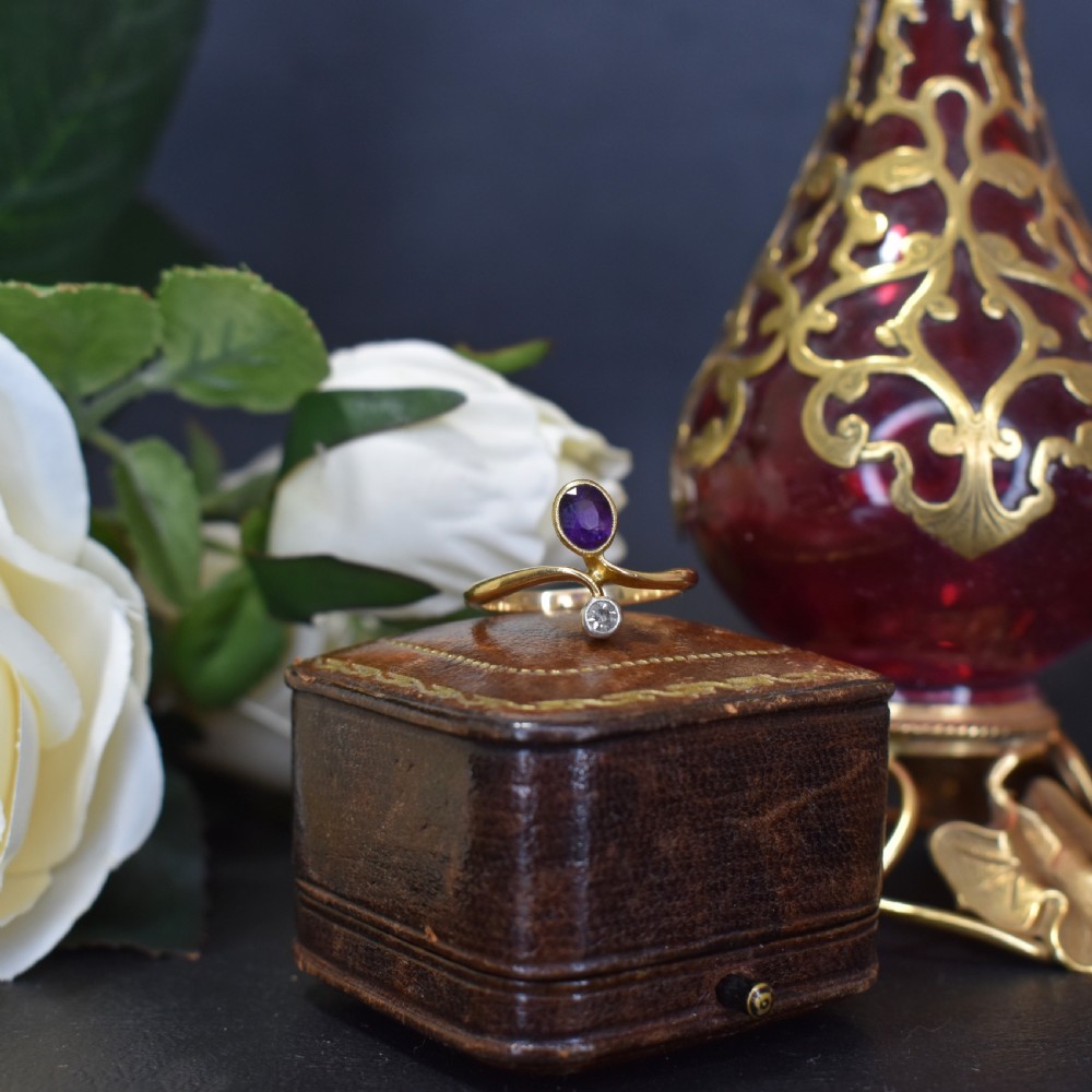 An Art Nouveau 18ct yellow gold ring featuring an oval bezel-set purple amethyst gemstone with a sweeping band and a small drop-set diamond accent. View IV