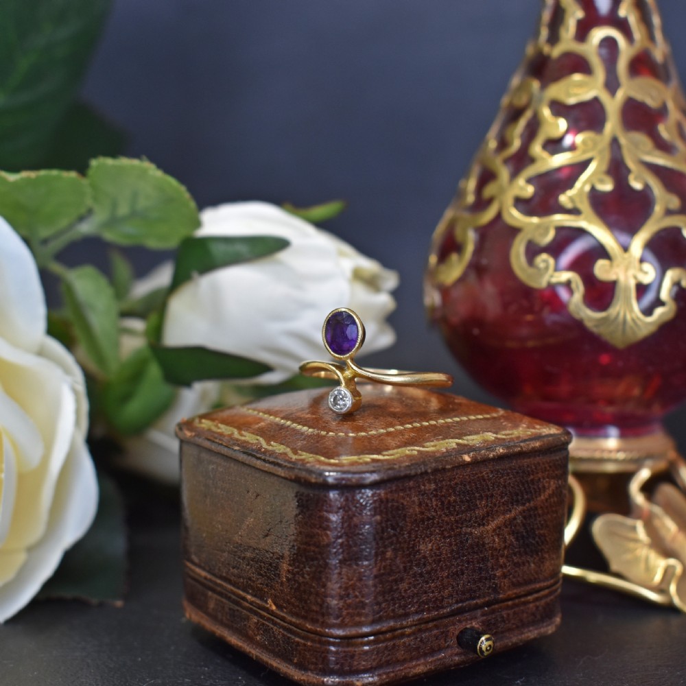 An Art Nouveau 18ct yellow gold ring featuring an oval bezel-set purple amethyst gemstone with a sweeping band and a small drop-set diamond accent. View V