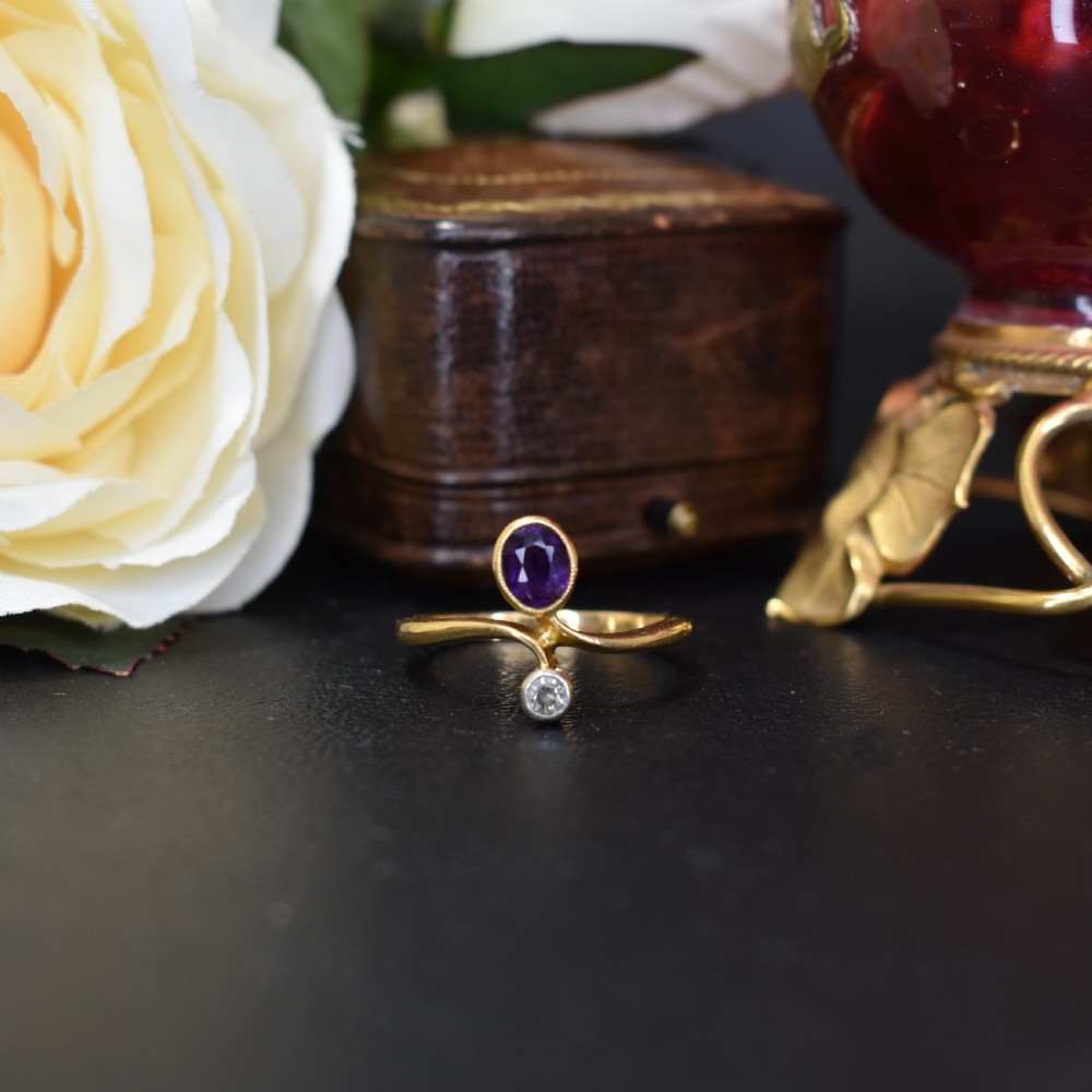 An Art Nouveau 18ct yellow gold ring featuring an oval bezel-set purple amethyst gemstone with a sweeping band and a small drop-set diamond accent. View VIII
