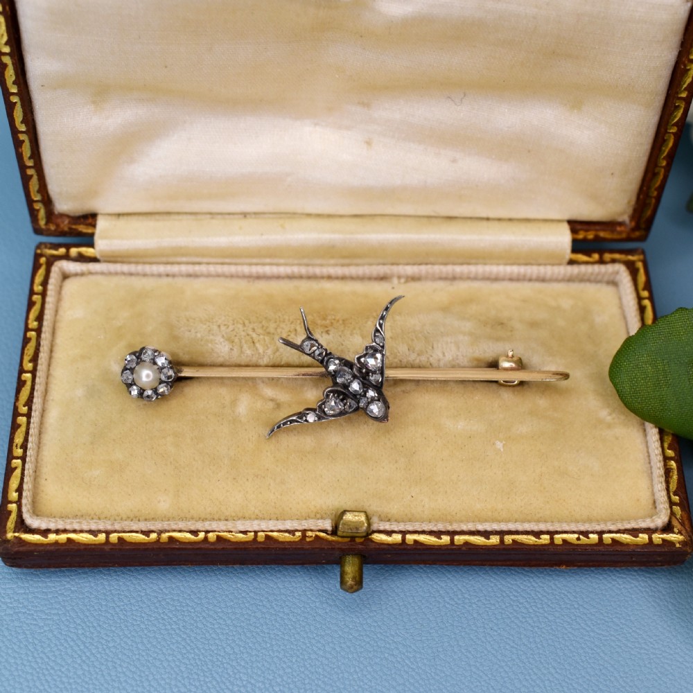 An Edwardian 15ct gold bar brooch featuring a silver-topped swallow in flight set with rose-cut diamonds beside a pearl cluster. View V