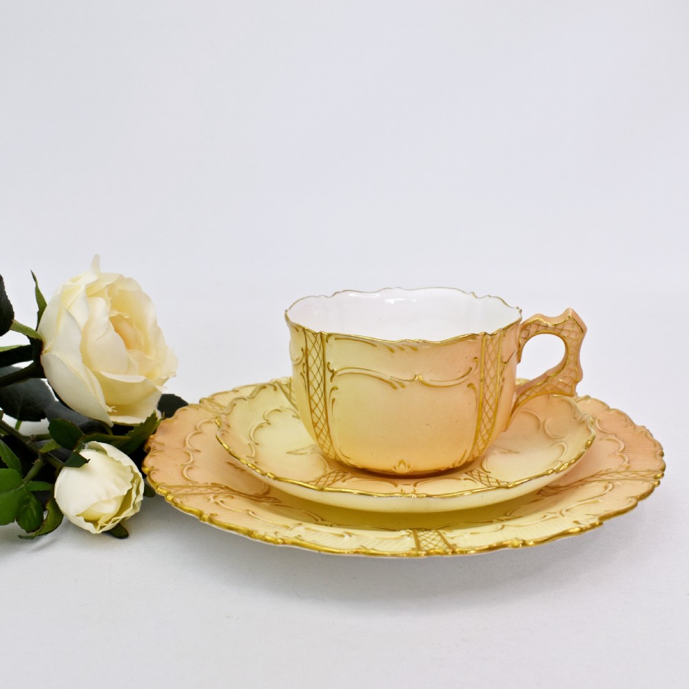 An Edwardian Royal Worcester tea trio featuring a teacup, saucer, and side plate rendered in gradient blush ivory porcelain with gilded scalloped edges.