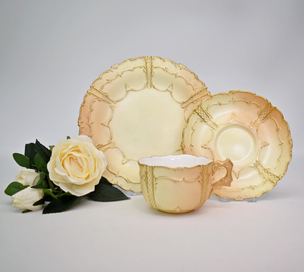 An Edwardian Royal Worcester tea trio featuring a teacup, saucer, and side plate rendered in gradient blush ivory porcelain with gilded scalloped edges. View II