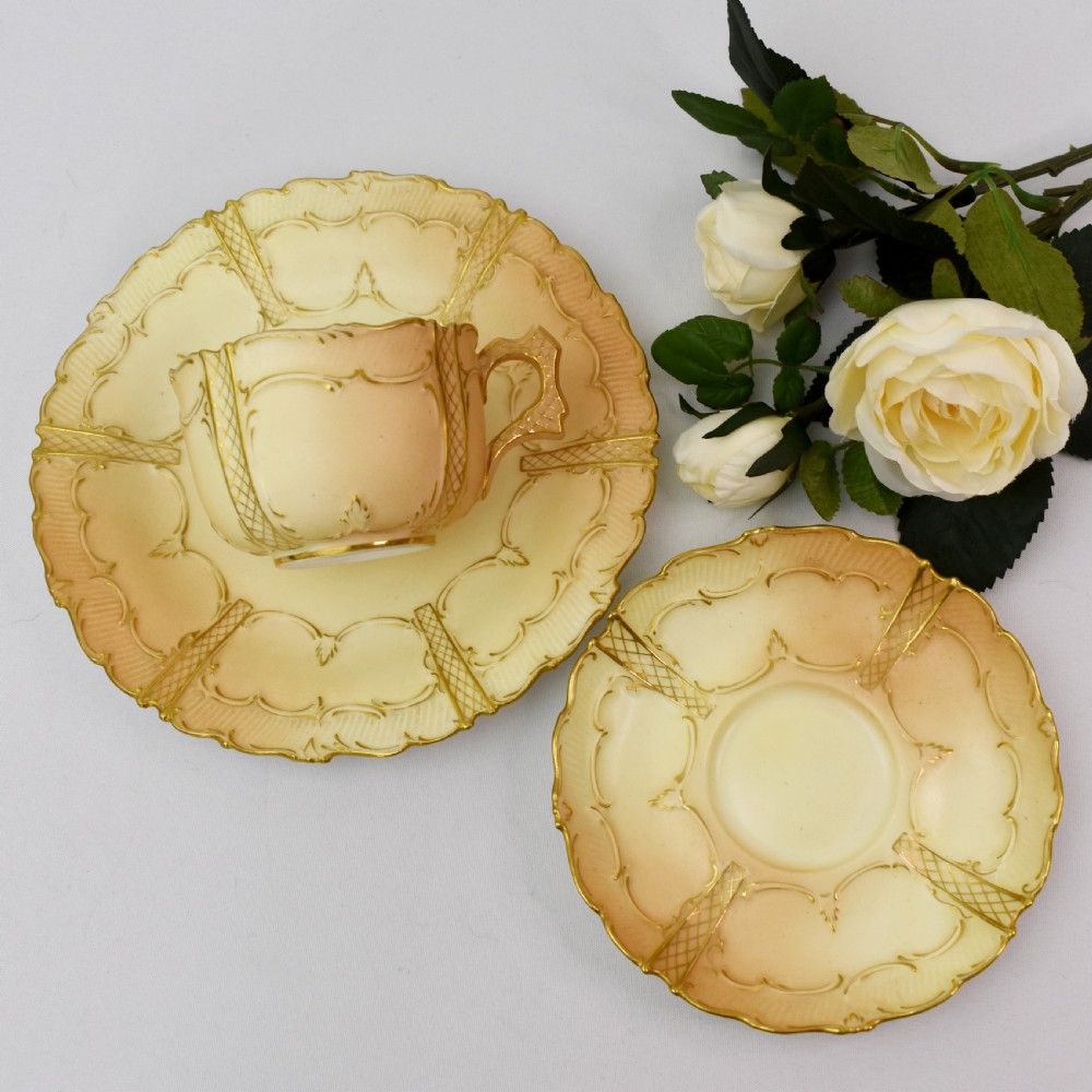 An Edwardian Royal Worcester tea trio featuring a teacup, saucer, and side plate rendered in gradient blush ivory porcelain with gilded scalloped edges. View V