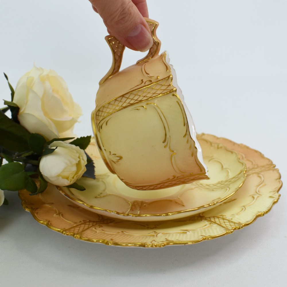 An Edwardian Royal Worcester tea trio featuring a teacup, saucer, and side plate rendered in gradient blush ivory porcelain with gilded scalloped edges. View VI