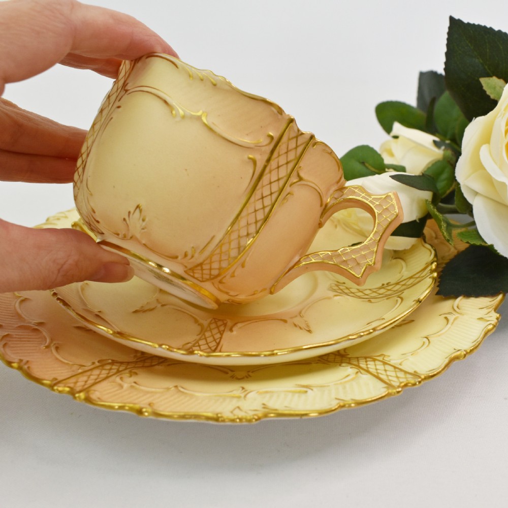 An Edwardian Royal Worcester tea trio featuring a teacup, saucer, and side plate rendered in gradient blush ivory porcelain with gilded scalloped edges. View VII
