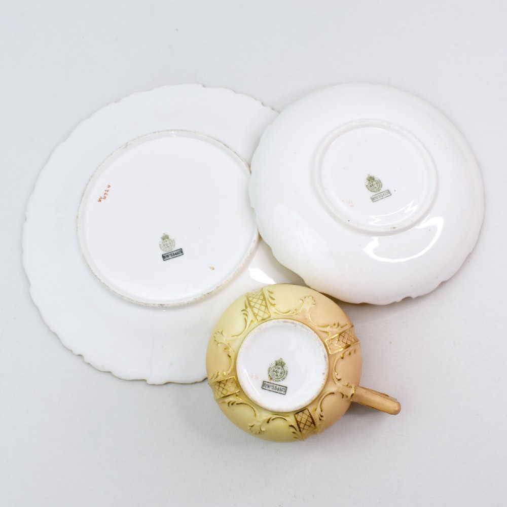 An Edwardian Royal Worcester tea trio featuring a teacup, saucer, and side plate rendered in gradient blush ivory porcelain with gilded scalloped edges. View X