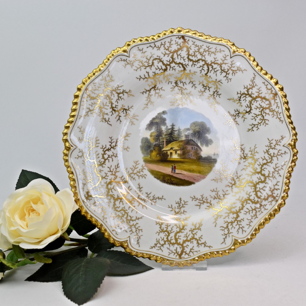 A Regency porcelain dessert plate featuring a hand-painted central landscape surrounded by intricate gold gilt seaweed patterns on a scalloped white border.