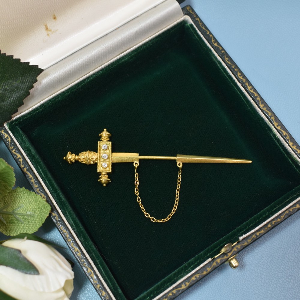 A Victorian 18 carat gold jabot pin crafted in the shape of a sword, featuring three old-cut diamonds set along the guard, cannetille wirework, and a safety chain.