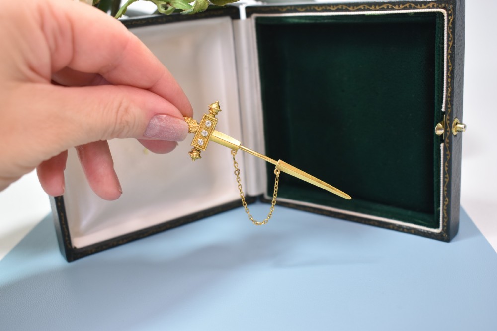 A Victorian 18 carat gold jabot pin crafted in the shape of a sword, featuring three old-cut diamonds set along the guard, cannetille wirework, and a safety chain. View IV