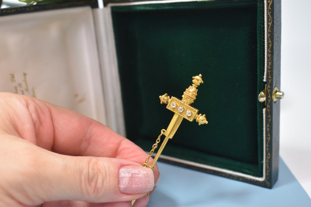 A Victorian 18 carat gold jabot pin crafted in the shape of a sword, featuring three old-cut diamonds set along the guard, cannetille wirework, and a safety chain. View V