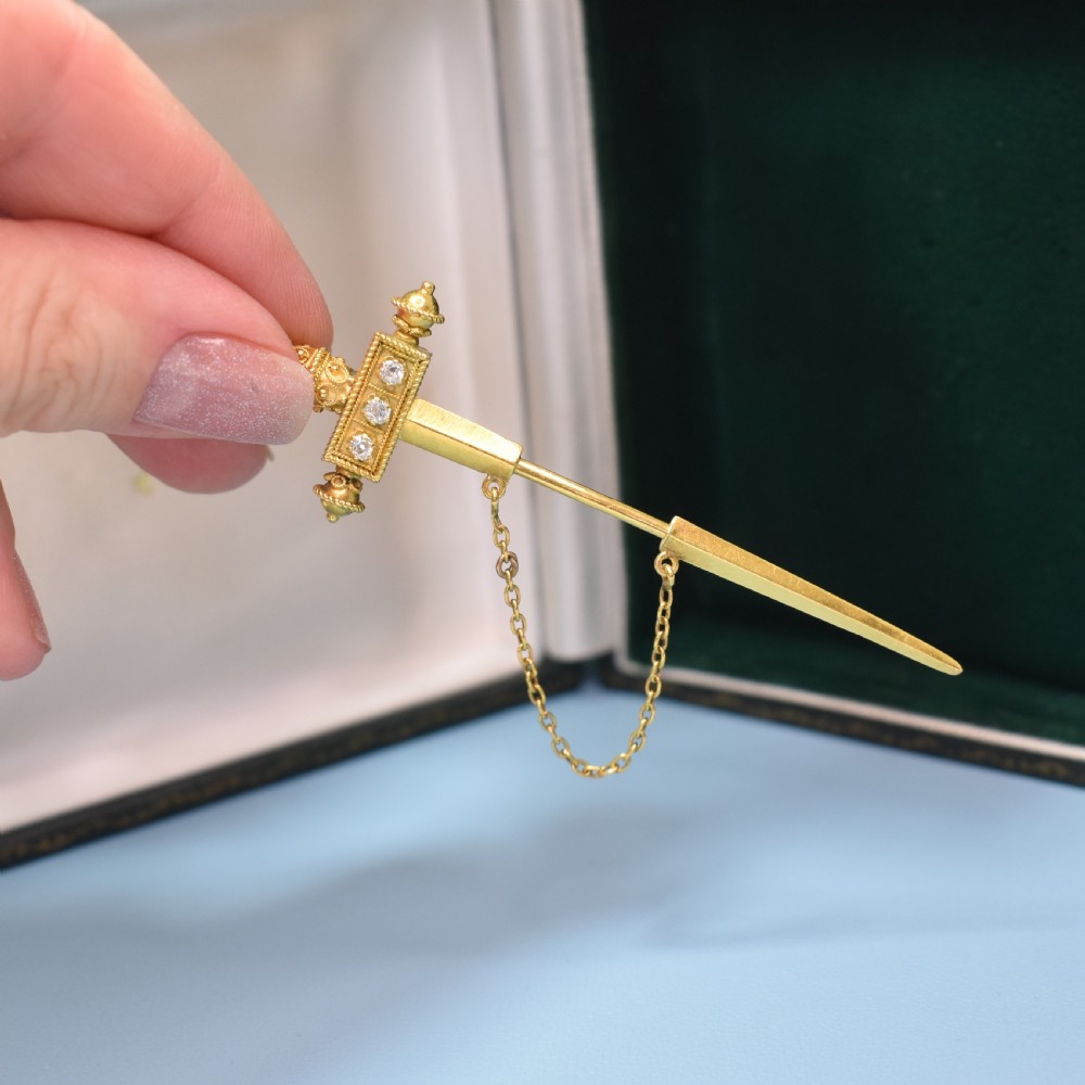 A Victorian 18 carat gold jabot pin crafted in the shape of a sword, featuring three old-cut diamonds set along the guard, cannetille wirework, and a safety chain. View VI