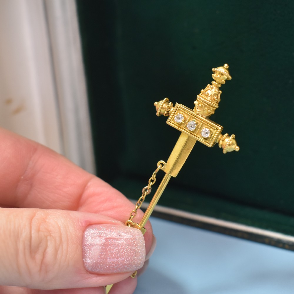 A Victorian 18 carat gold jabot pin crafted in the shape of a sword, featuring three old-cut diamonds set along the guard, cannetille wirework, and a safety chain. View VII