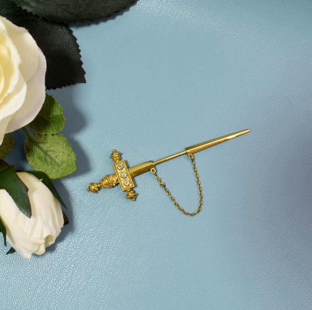 A Victorian 18 carat gold jabot pin crafted in the shape of a sword, featuring three old-cut diamonds set along the guard, cannetille wirework, and a safety chain. View VIII