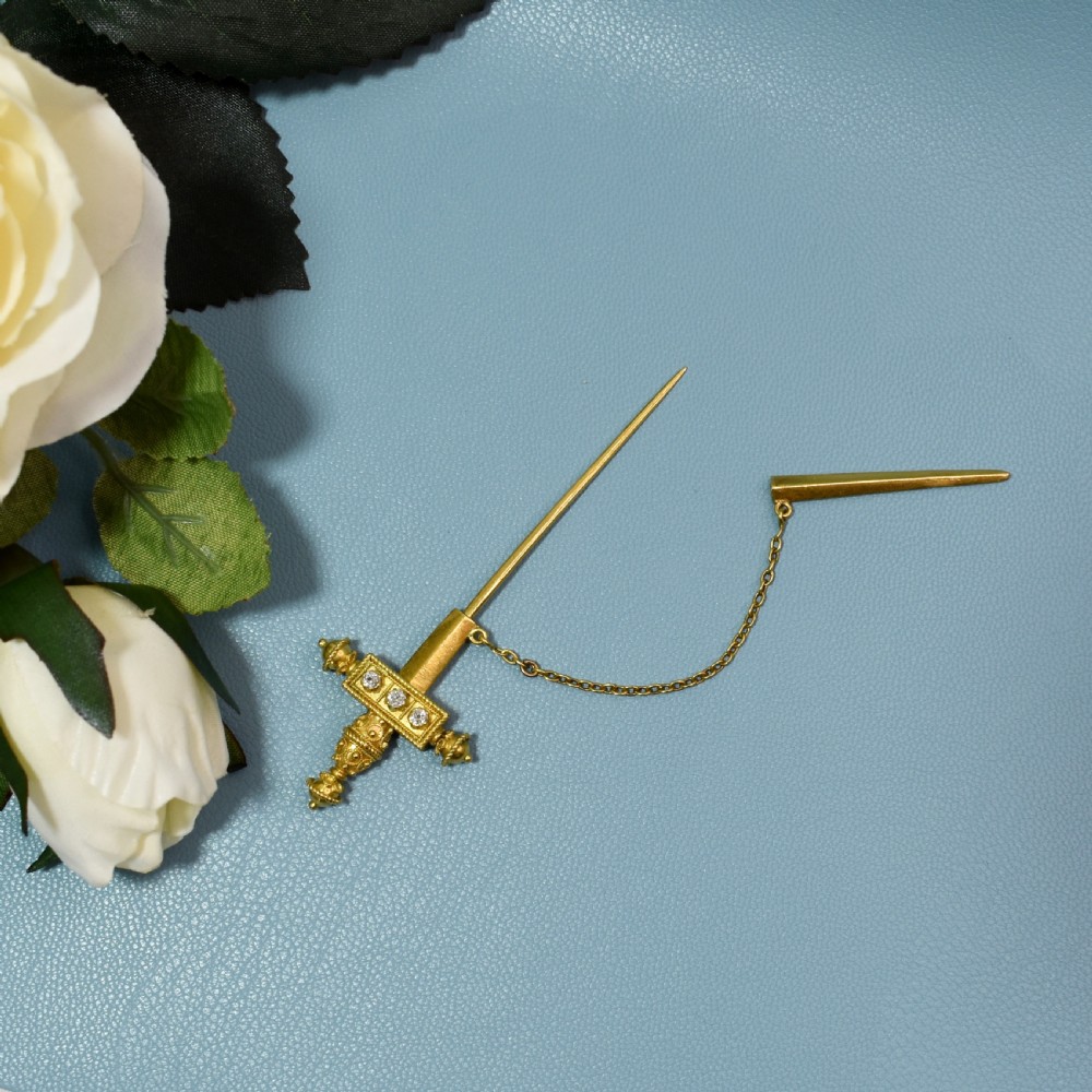 A Victorian 18 carat gold jabot pin crafted in the shape of a sword, featuring three old-cut diamonds set along the guard, cannetille wirework, and a safety chain. View IX
