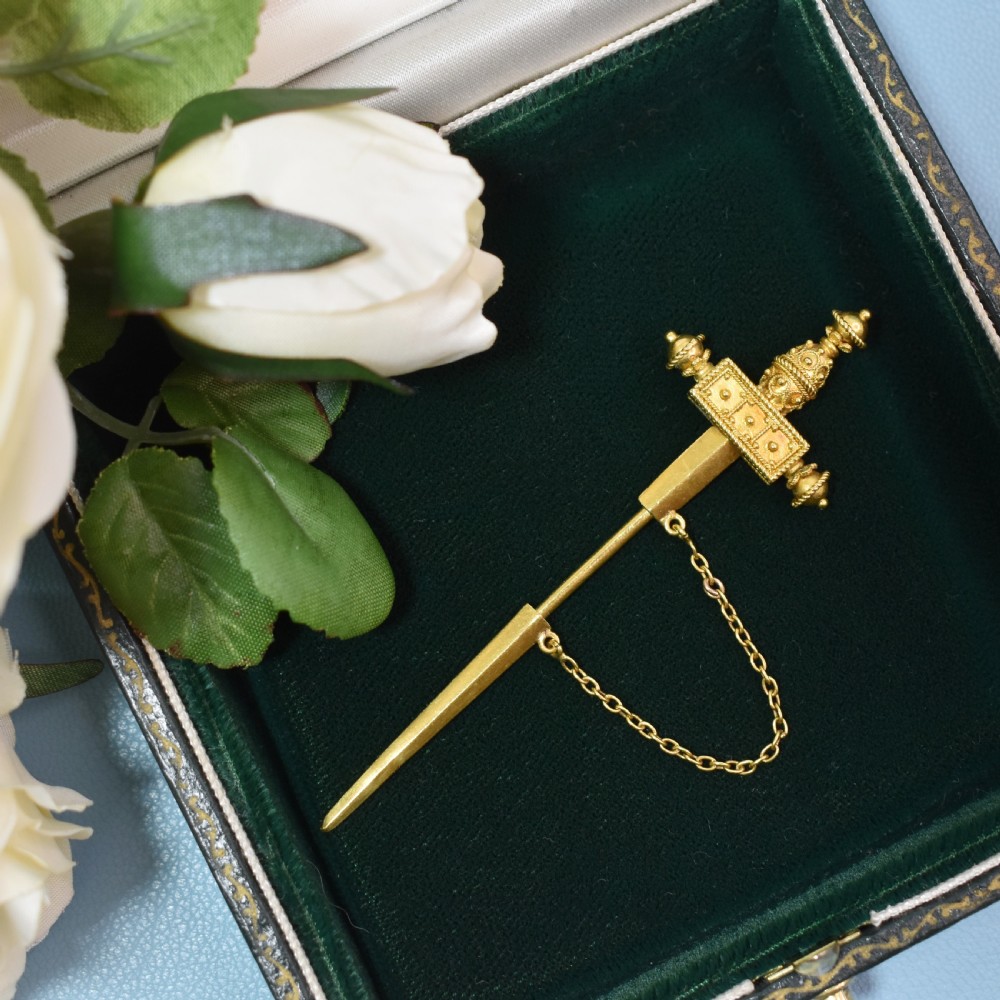 A Victorian 18 carat gold jabot pin crafted in the shape of a sword, featuring three old-cut diamonds set along the guard, cannetille wirework, and a safety chain. View X