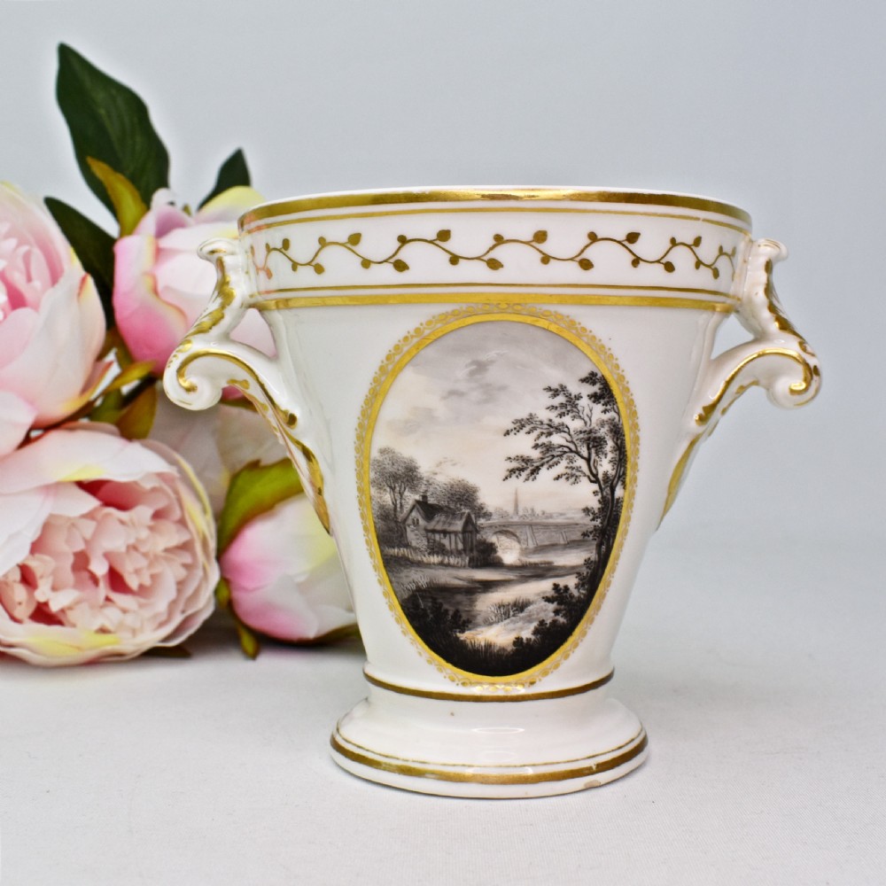 A white porcelain urn featuring two gilded scroll handles, a gilded rim with a vine motif, and an oval medallion painted with a monochrome landscape.