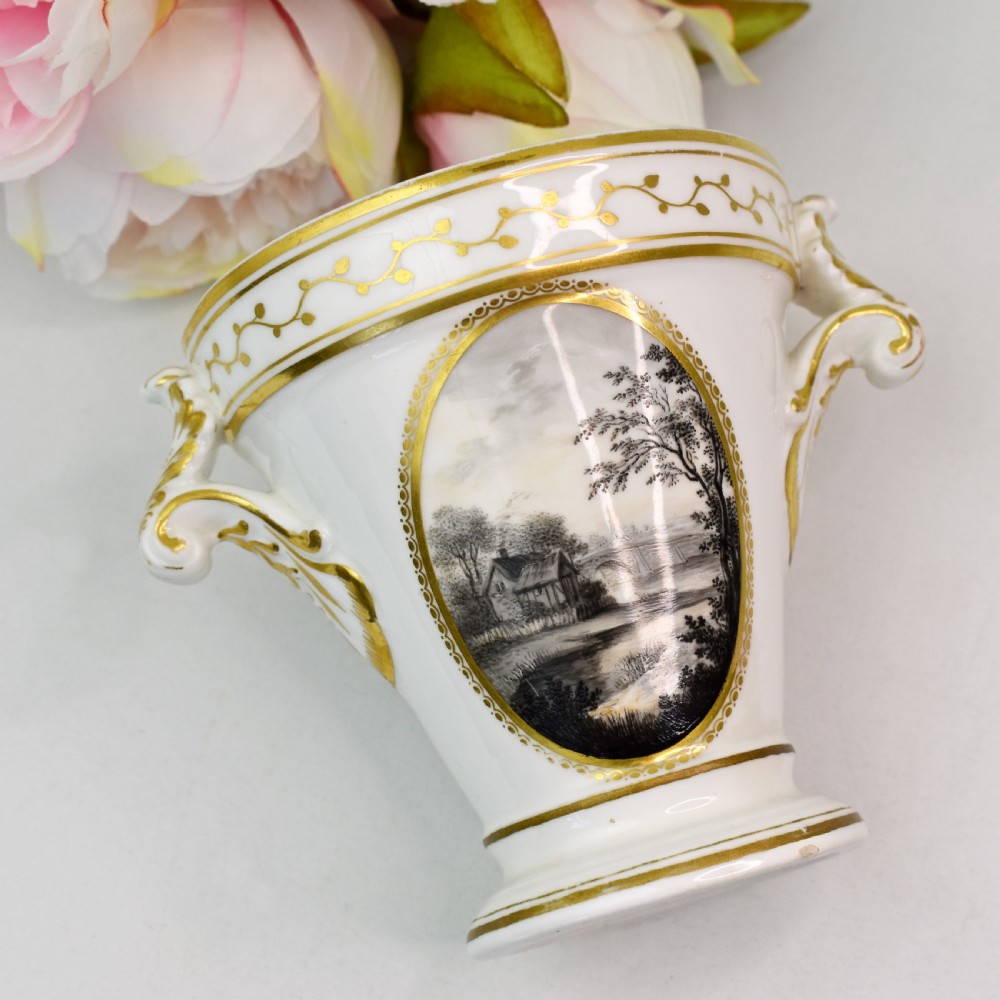 A white porcelain urn featuring two gilded scroll handles, a gilded rim with a vine motif, and an oval medallion painted with a monochrome landscape. View III