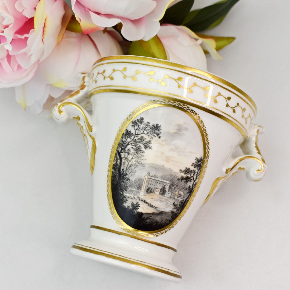 A white porcelain urn featuring two gilded scroll handles, a gilded rim with a vine motif, and an oval medallion painted with a monochrome landscape. View IV