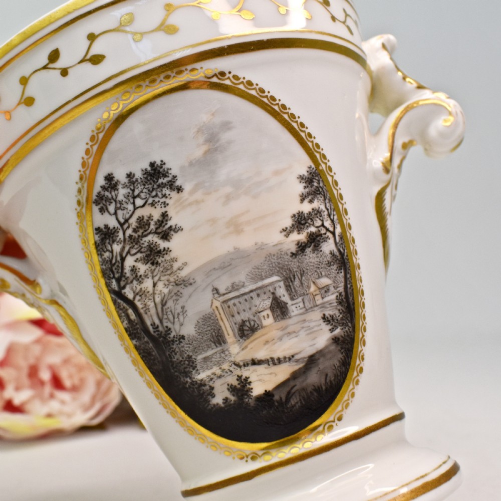 A white porcelain urn featuring two gilded scroll handles, a gilded rim with a vine motif, and an oval medallion painted with a monochrome landscape. View V