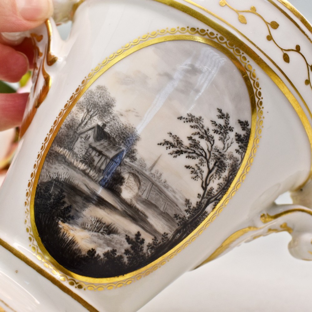 A white porcelain urn featuring two gilded scroll handles, a gilded rim with a vine motif, and an oval medallion painted with a monochrome landscape. View VI