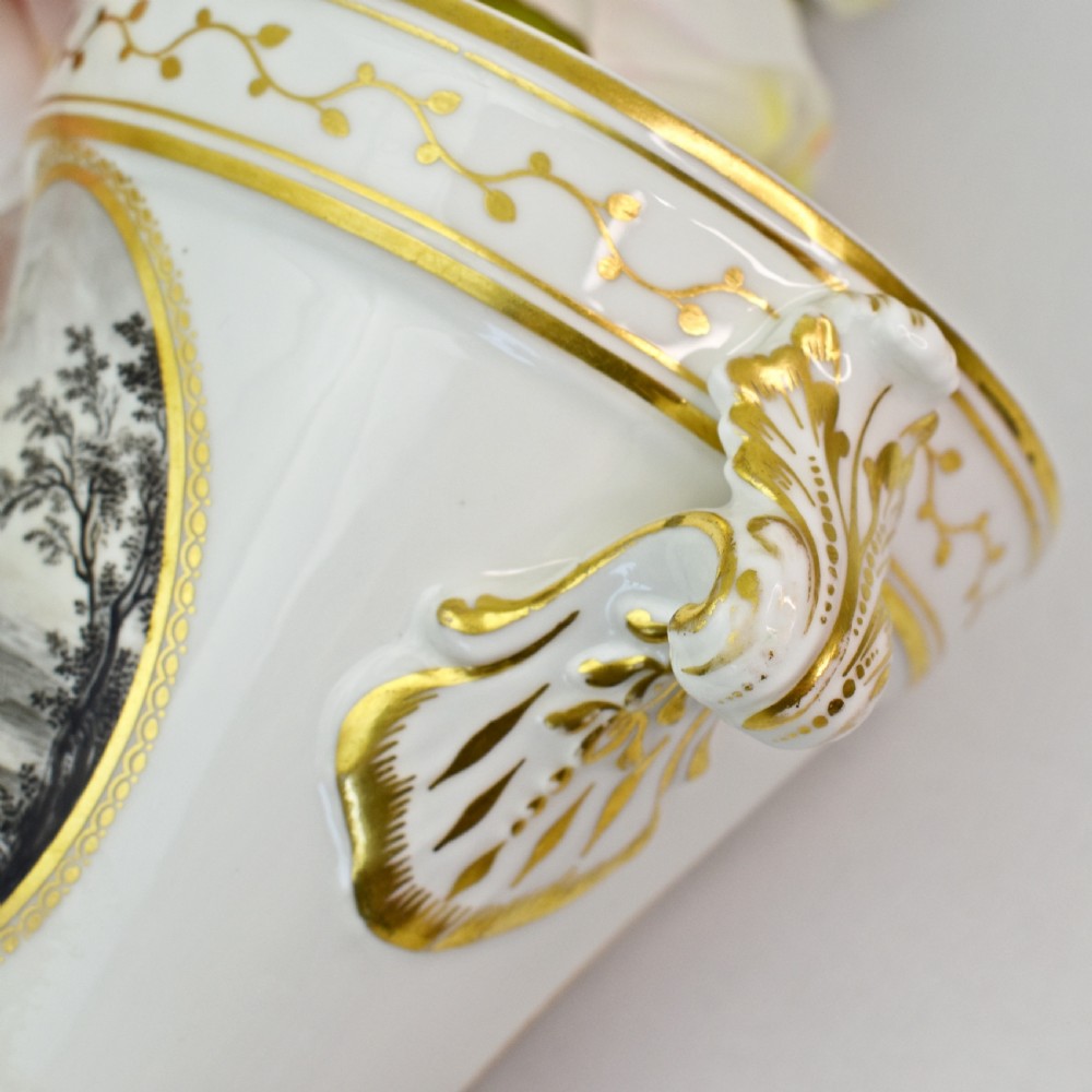 A white porcelain urn featuring two gilded scroll handles, a gilded rim with a vine motif, and an oval medallion painted with a monochrome landscape. View VII