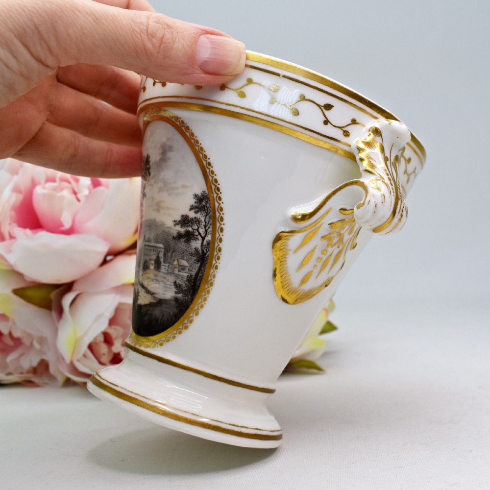 A white porcelain urn featuring two gilded scroll handles, a gilded rim with a vine motif, and an oval medallion painted with a monochrome landscape. View VIII