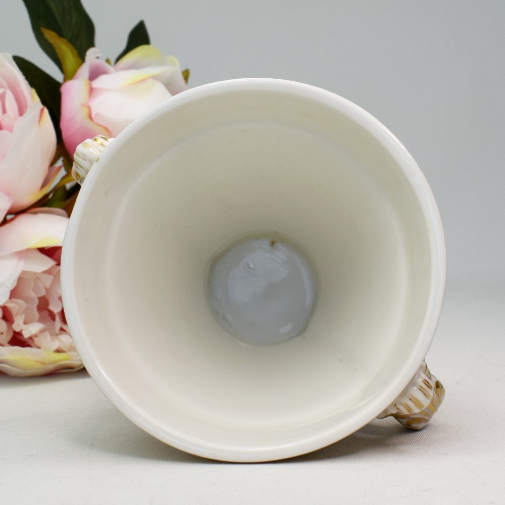 A white porcelain urn featuring two gilded scroll handles, a gilded rim with a vine motif, and an oval medallion painted with a monochrome landscape. View IX