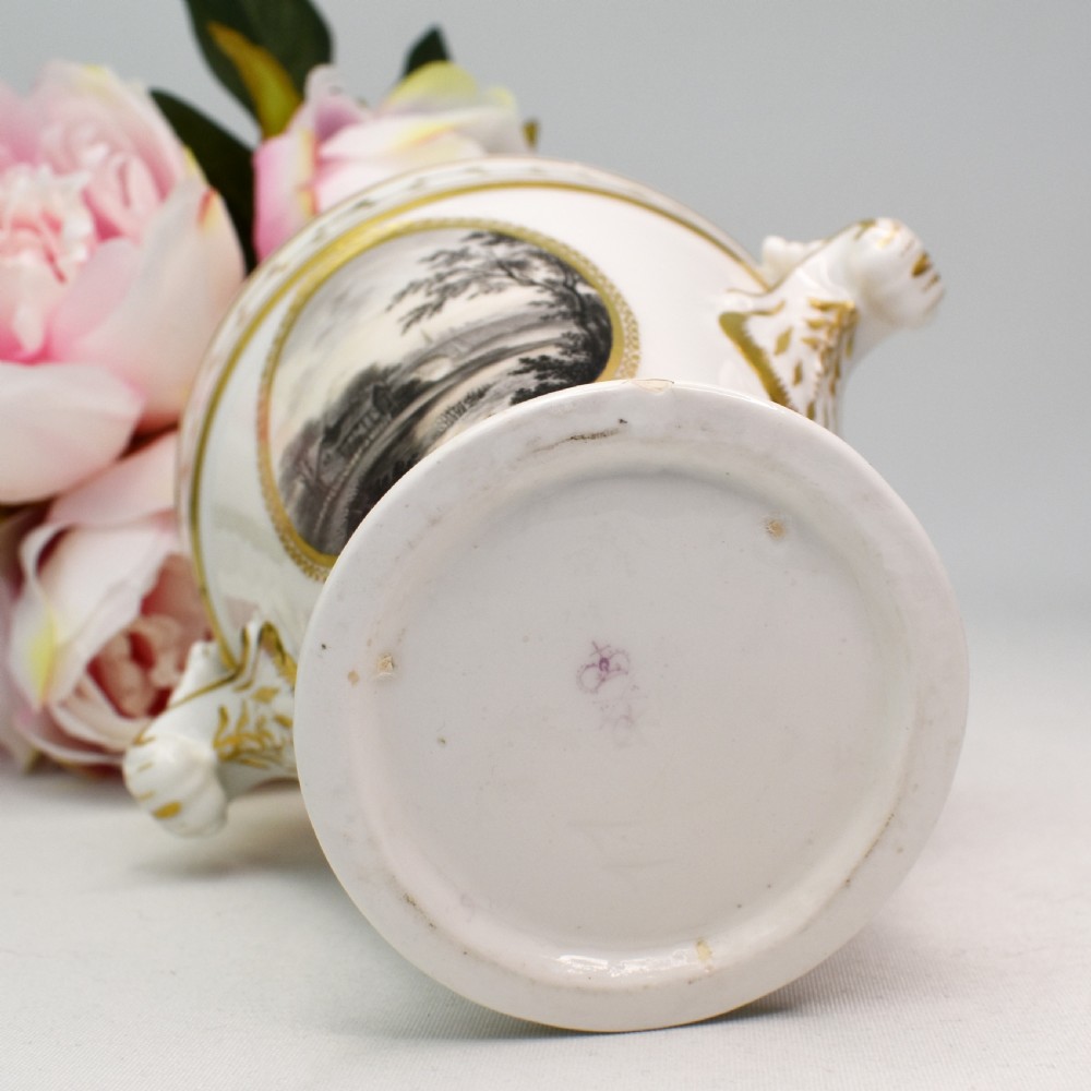A white porcelain urn featuring two gilded scroll handles, a gilded rim with a vine motif, and an oval medallion painted with a monochrome landscape. View X