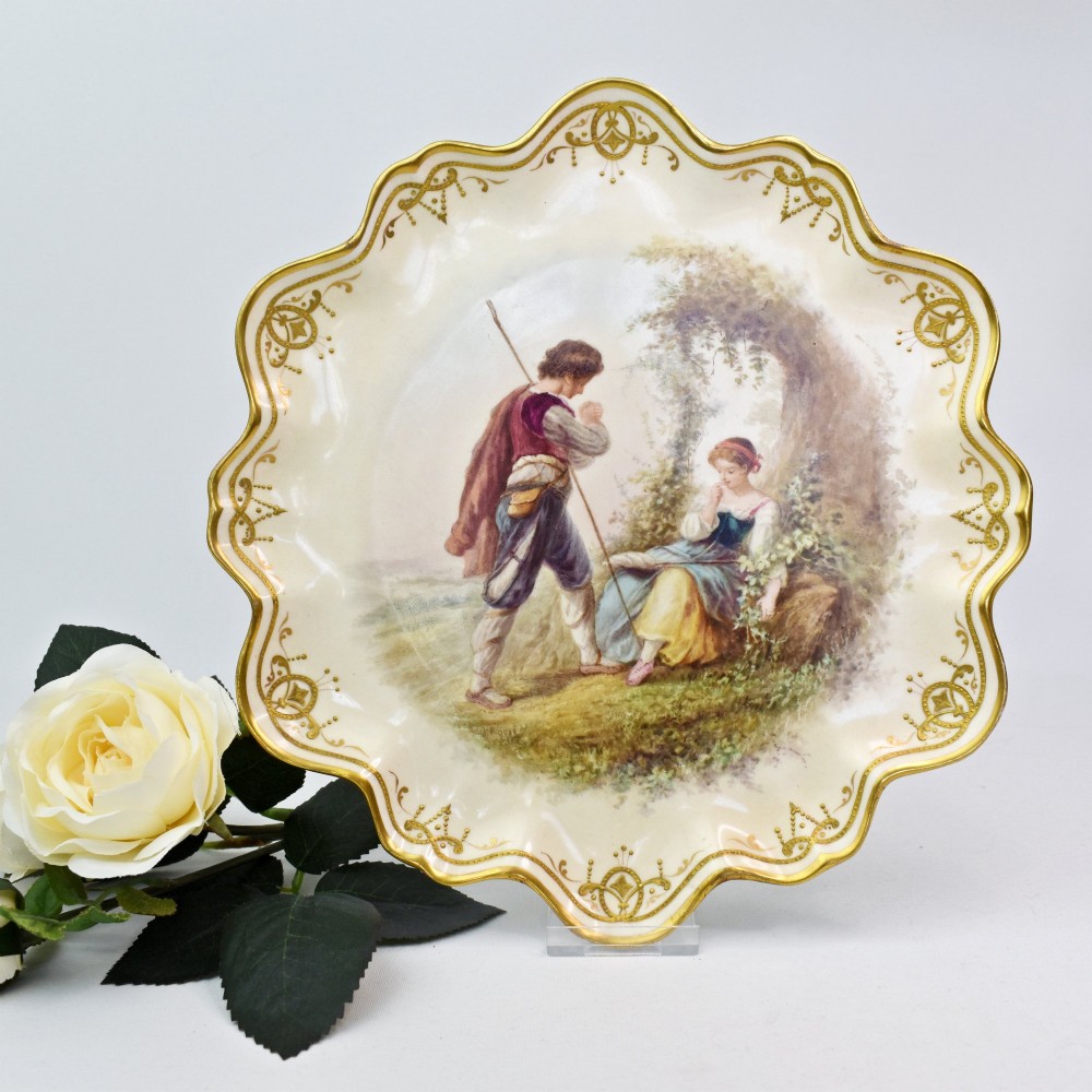 A circular Victorian porcelain cabinet plate featuring a hand-painted romantic pastoral scene enclosed by a lobed rim decorated with gold gilding.