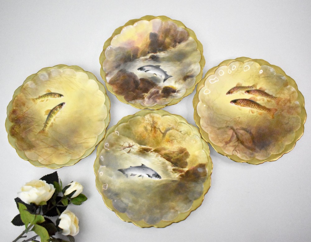 A set of four circular porcelain cabinet plates with scalloped gold rims, each featuring a central hand-painted scene of fish swimming in natural river settings.